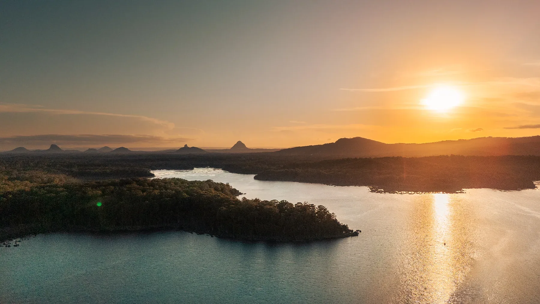 Golden sunlight reflected across a lake beneath the silhouettes of the Glasshouse Mountains